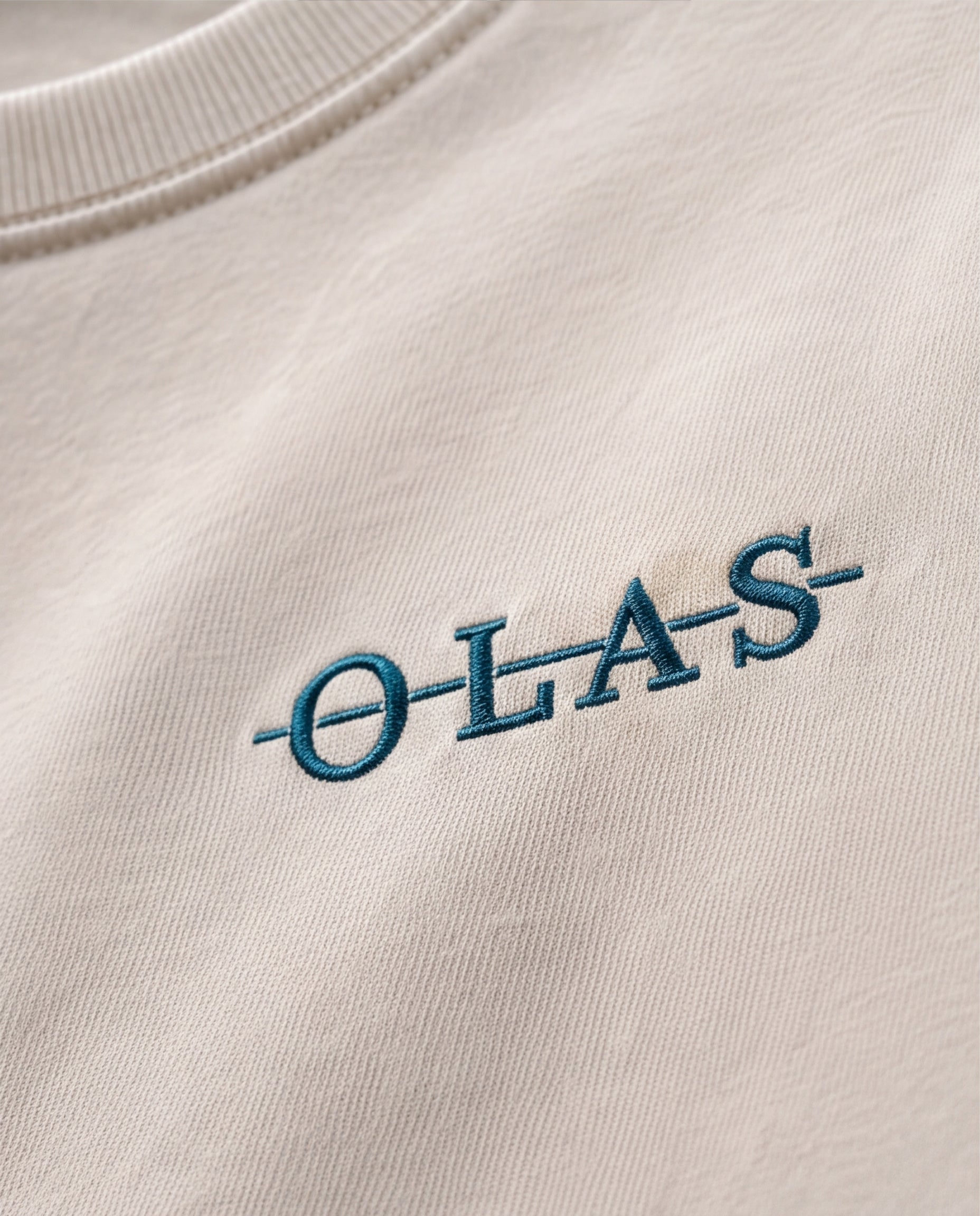Close-up of the embroidered crossed Olas chest mark on cream heavyweight cotton